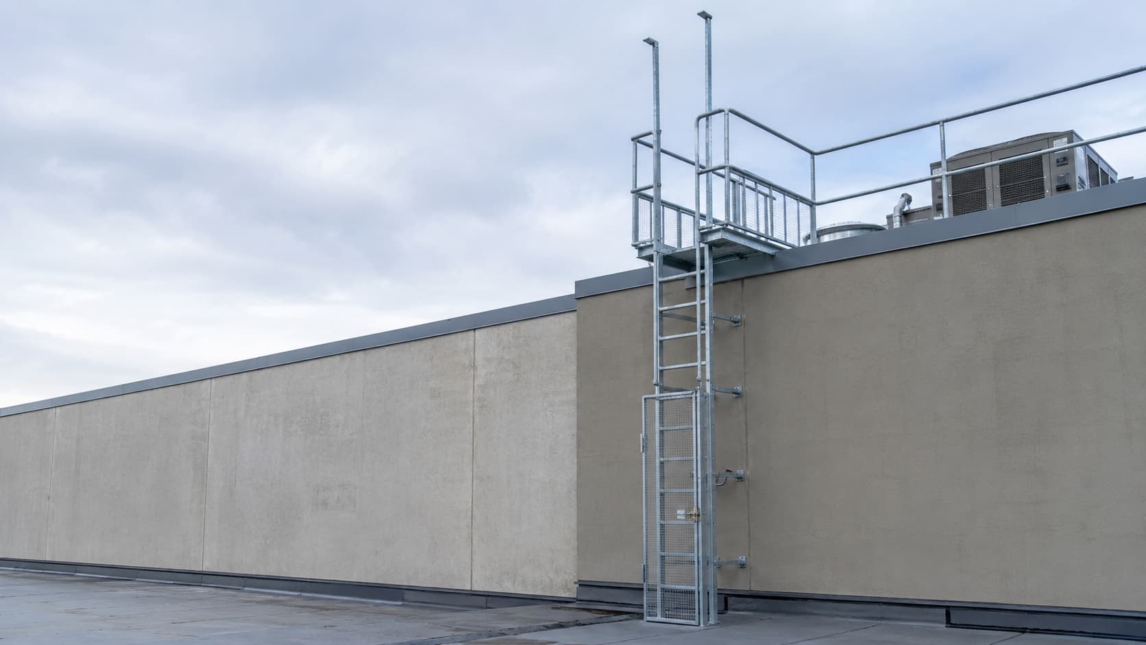 Galvanized fixed roof access ladder with a safety cage and guardrail on a concrete building wall