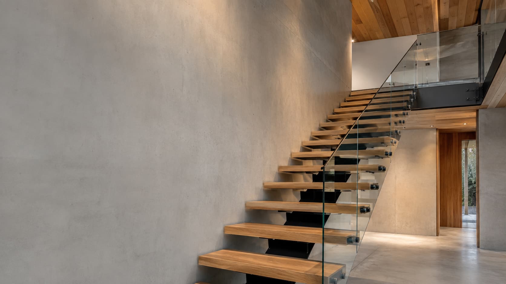 Floating oak stair treads cantilevered from a black steel spine with a frameless glass railing in a concrete-walled home