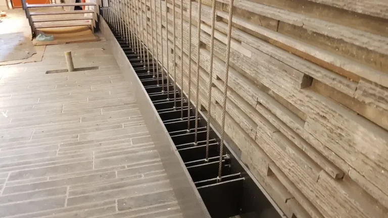 Vertical steel wire ropes tensioned along a floor edge channel beside a stone clad wall