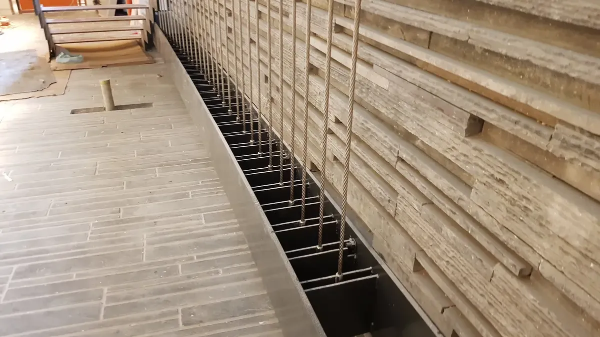 Vertical steel wire ropes tensioned along a floor edge channel beside a stone clad wall